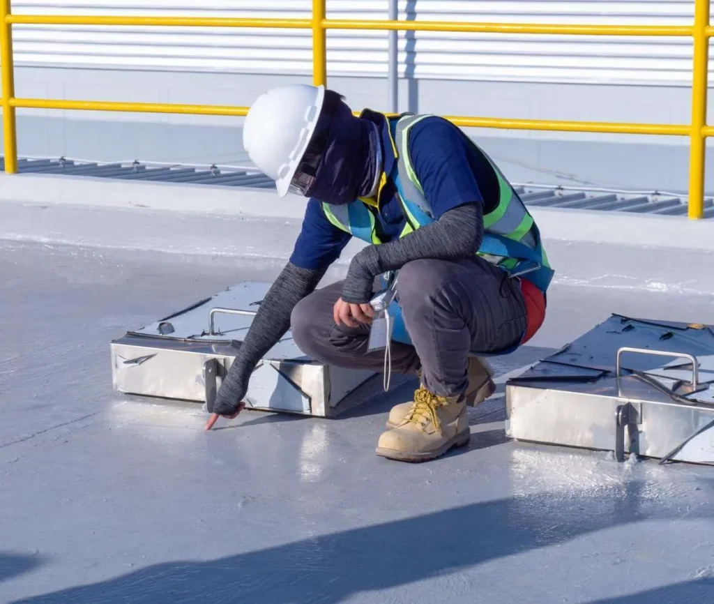 Roofing technician in a hard hat inspecting a freshly coated commercial roof around steel curbs