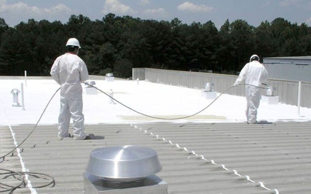 Two workers in white protective suits spray-applying white coating to a metal commercial roof